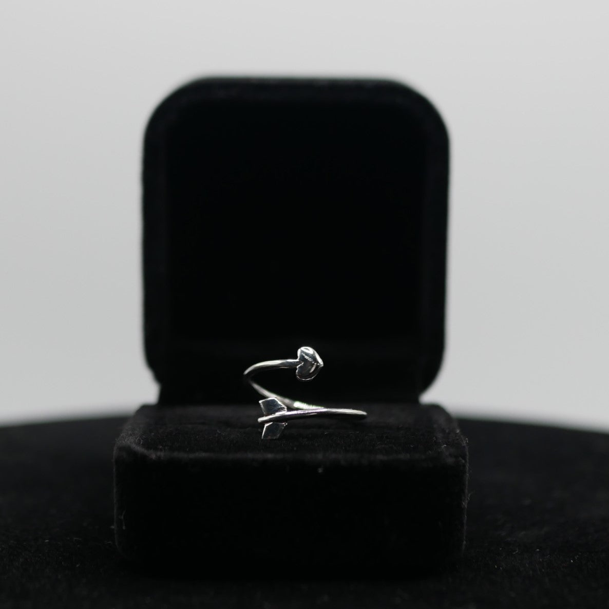 A silver ring that look like an arrow with a heart in a black jewelry box