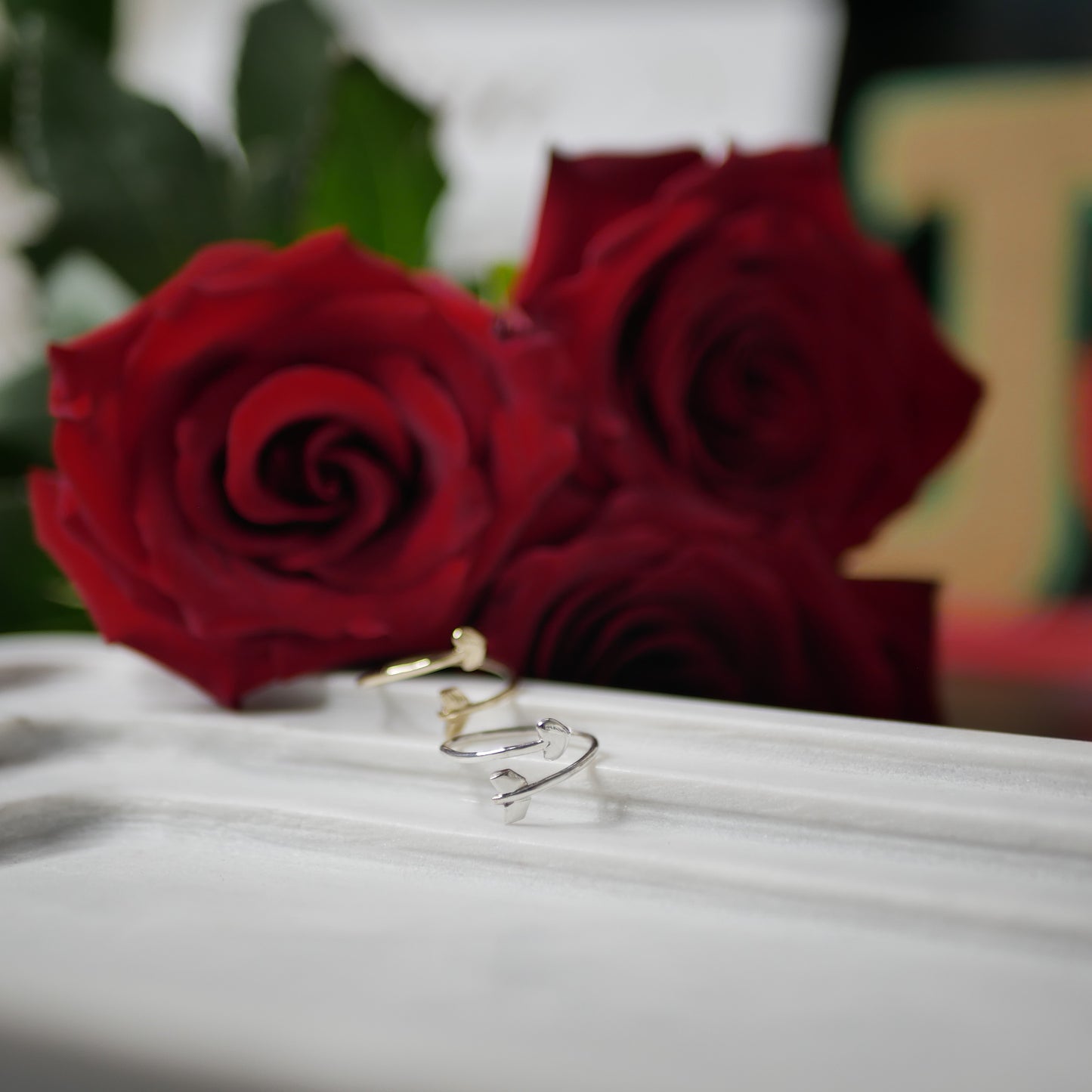 A silver heart arrow ring displayed in front of another ring and three red roses
