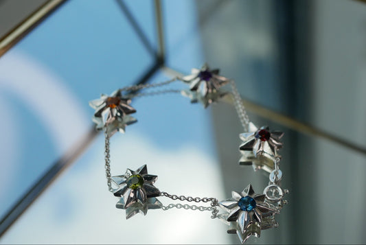 A silver-toned chain bracelet featuring five starburst-shaped charms, each set with a different colored gemstone (blue, purple, orange, green, and clear), photographed hanging against a reflective glass surface.