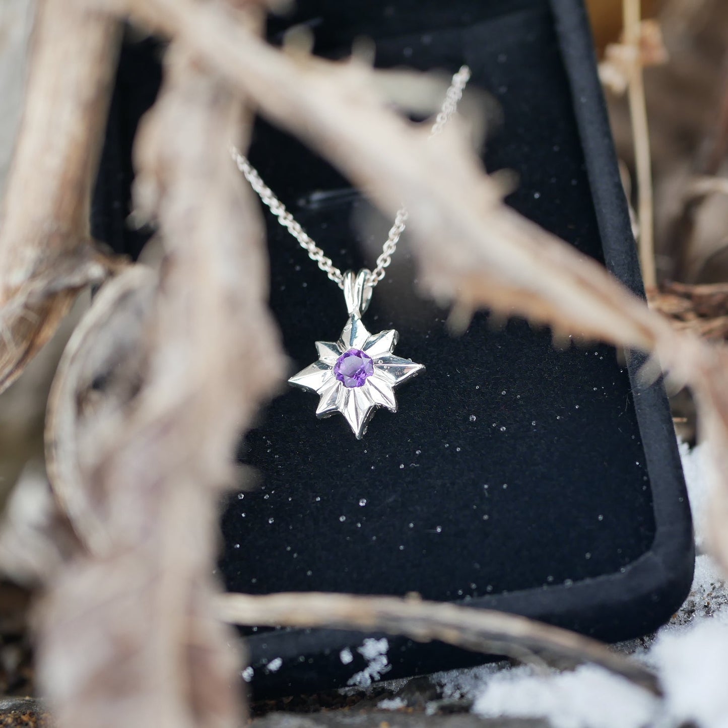 Star-shaped pendant with a purple gemstone on a black stand, surrounded by natural elements.