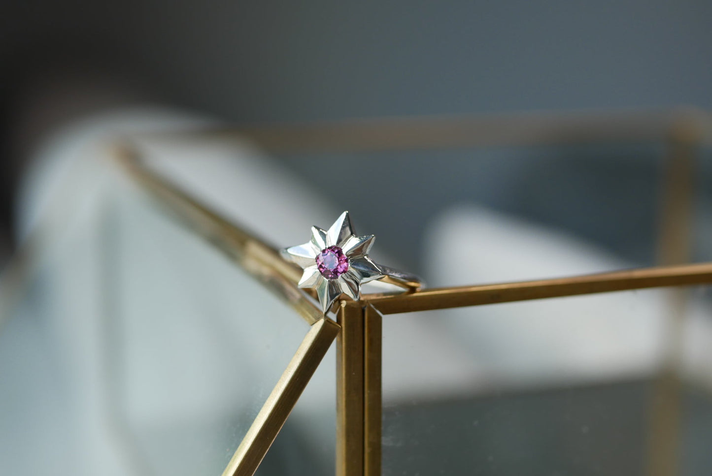 A silver starburst ring with a small pink gemstone in the center, resting on the corner of a gold-framed glass display case.